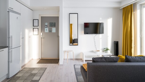 A modern living room with a grey sofa, a flat-screen TV mounted on the wall, and a dining area with a white table and chairs.