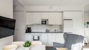 A modern kitchen with white cabinets, a microwave, and a refrigerator. There is a gray sofa and a white table with a bowl and plant.