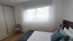 A bedroom with a bed, a chair, a window with sheer curtains, and a nightstand with a lamp. - Atlantic Valley - Furnas (Furnas)
