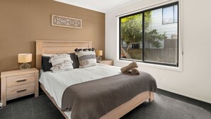A bedroom with a large bed, wooden headboard, two bedside tables, a window with a view of greenery, and a decorative wall piece. - Sunderland Retreat in Sunderland Bay (Sunderland Bay)