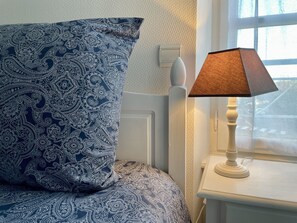 Une chambre avec un couvre-lit bleu et blanc à motifs, une table de chevet blanche avec une lampe, et une fenêtre donnant sur l’extérieur.
