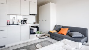 A modern kitchen with white cabinets, a built-in oven, and a coffee maker. There is a grey sofa with orange and grey cushions in the living area. - Hiisi Homes Nokia (Nokia)