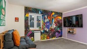 A room with a colorful graffiti wall, a grey sofa with orange cushions, a wooden table, a radiator, and a television on the wall. - Ash - Central, Indoor Playground, Garden, Switch & Stream, SummerCard (Winterberg)