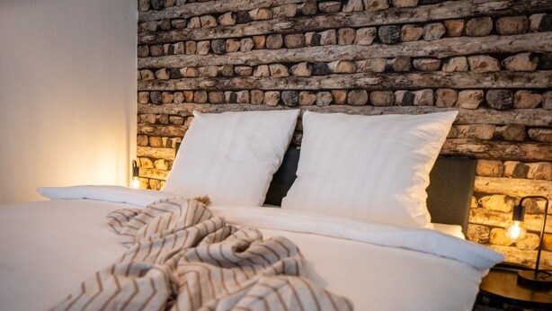 A bed with white linens and striped bedding, a stone wall behind, and a nightstand with a lamp. - Ash - Central, Indoor Playground, Garden, Switch & Stream, SummerCard (Winterberg)