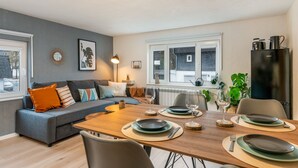 A modern living room with a grey sofa, wooden dining table set for four, and a black refrigerator. - Ash - Central, Indoor Playground, Garden, Switch & Stream, SummerCard (Winterberg)