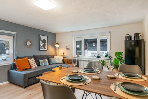 A modern living room with a grey sofa, wooden dining table set for four, and a black refrigerator.