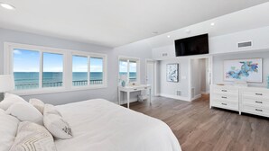 Ein Schlafzimmer mit einem großen Bett, einem Schreibtisch und einem Fernseher. Es gibt Fenster mit Blick aufs Meer.