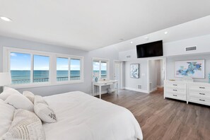 A bedroom with a large bed, a desk, and a TV. There are windows with a view of the ocean.