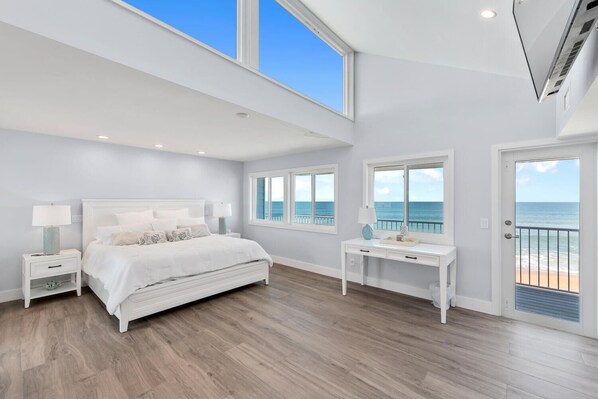 A spacious bedroom with a large bed, two bedside tables, a desk, and a view of the ocean through large windows.