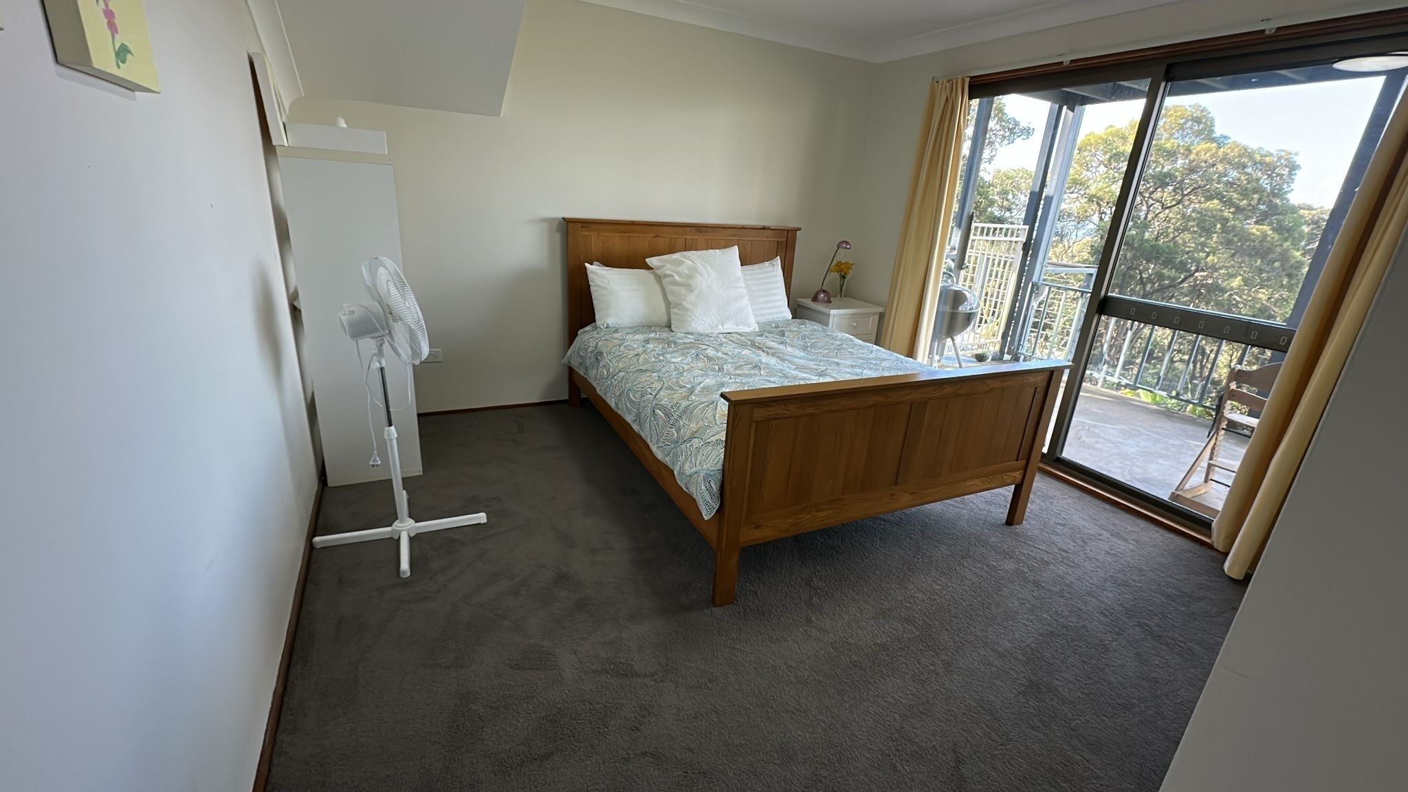Ein Schlafzimmer mit einem Holzbett, einem Standventilator und Blick ins Freie durch eine Schiebetür.