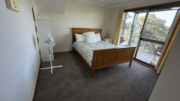 Ein Schlafzimmer mit einem Holzbett, einem Standventilator und Blick ins Freie durch eine Schiebetür.