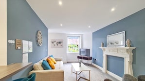 A modern living room with a sofa, coffee table, TV, fireplace, and a large window. - Margaret's Garden by Fortuna Property Portfolio, The Margaret's Garden (Cheltenham)