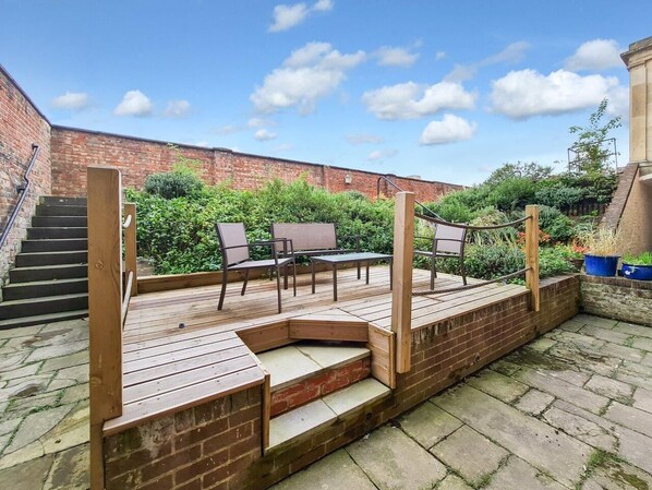 A wooden deck with outdoor seating and a brick wall in the background. - Margaret's Garden by Fortuna Property Portfolio, The Margaret's Garden (Cheltenham)