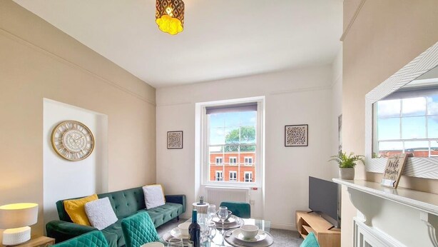 Uma sala de estar moderna com uma mesa de jantar de vidro posta para quatro pessoas, sofás estofados em tons de verde-água, uma janela grande com vista para um edifício de tijolos vermelhos e um relógio fixado na parede.