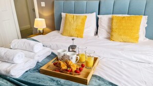 Cama bem arrumada com cabeceira azul, almofadas brancas e bandeja com croissants, morangos e suco.