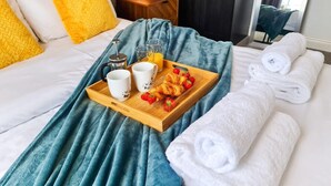 En snyggt bäddad säng med ett blått täcke, ett bricka med croissanter, jordgubbar och juice, och hoprullade vita handdukar.