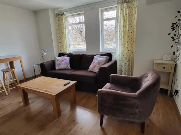 A living room with a wooden coffee table, a brown sofa with floral cushions, a brown armchair, and a wooden side table with a lamp.