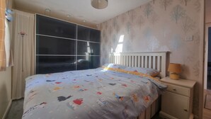 A bedroom with a bed, a nightstand, a lamp, a window with curtains, and a wall with a tree pattern.
