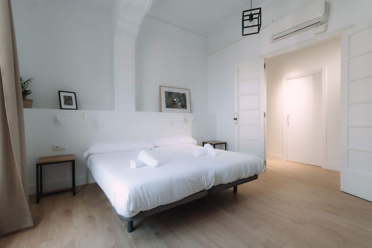 A modern bedroom with a large bed, bedside tables, a wall-mounted air conditioner, and a door leading to another room.