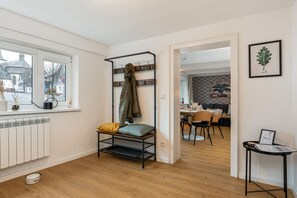 Um quarto com radiador, uma janela com vista para a rua, um cabideiro com um casaco, uma sala de jantar com cadeiras e uma mesa, e um quadro na parede.