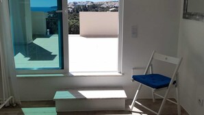 Una habitación con un gran ventanal corredero que da a un balcón, una silla blanca con cojín azul y una alfombra estampada en el suelo.