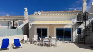 Zona de piscina con sillones, una mesa y un porche cubierto.