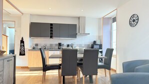 A modern kitchen with grey cabinets, a central island, and a dining area with black chairs. - Les Pensées (Saint-Vigor-le-Grand)