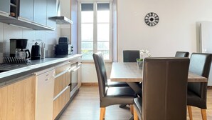 A modern kitchen with a built-in oven, a coffee maker, and a dishwasher. There is a dining table with chairs and a clock on the wall. - Les Pensées (Saint-Vigor-le-Grand)