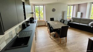 A modern kitchen with dark cabinets, a wooden dining table, and a black sofa. - Les Pensées (Saint-Vigor-le-Grand)