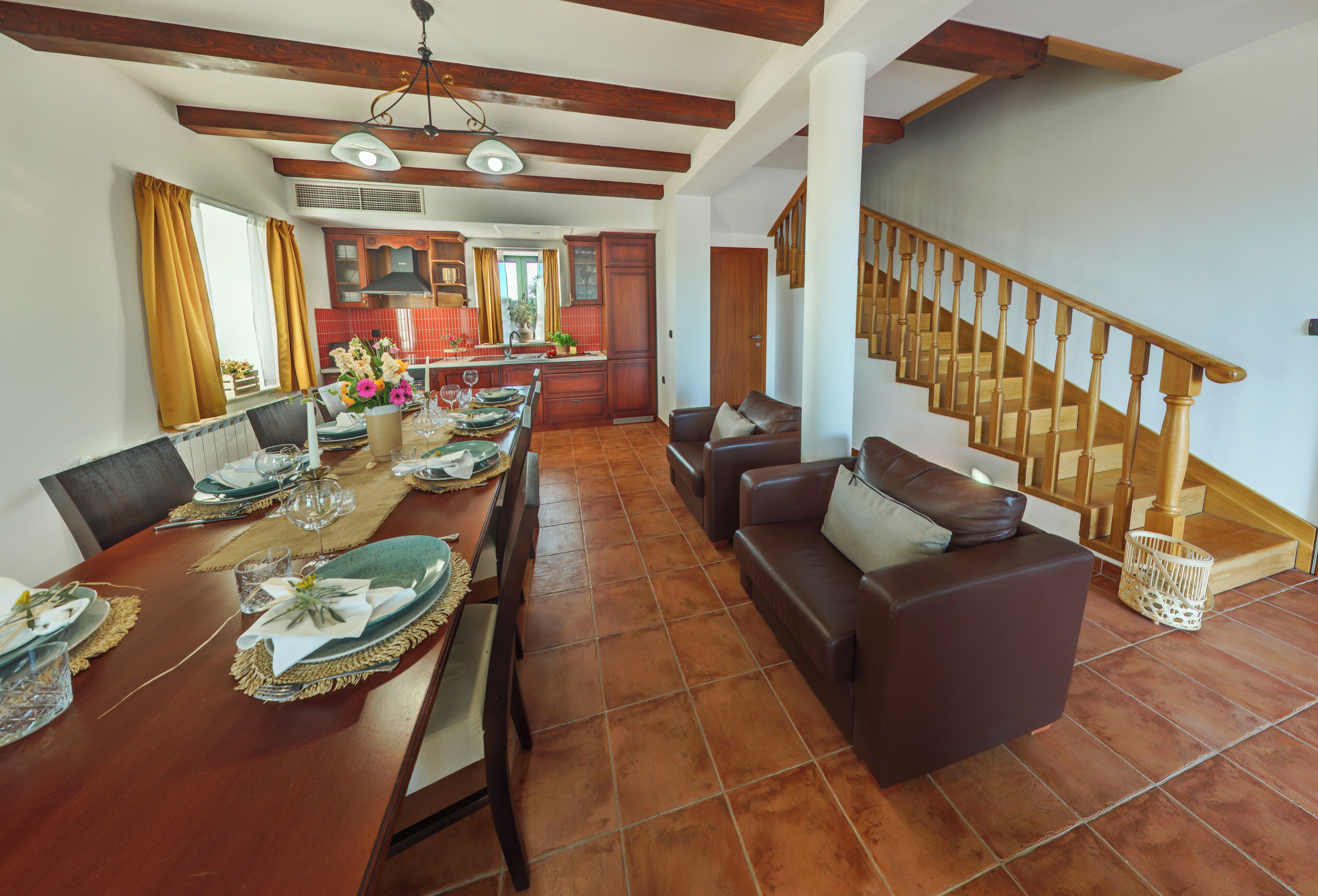 A dining area with a wooden table set for a meal, a brown leather sofa, and a staircase with wooden railings.