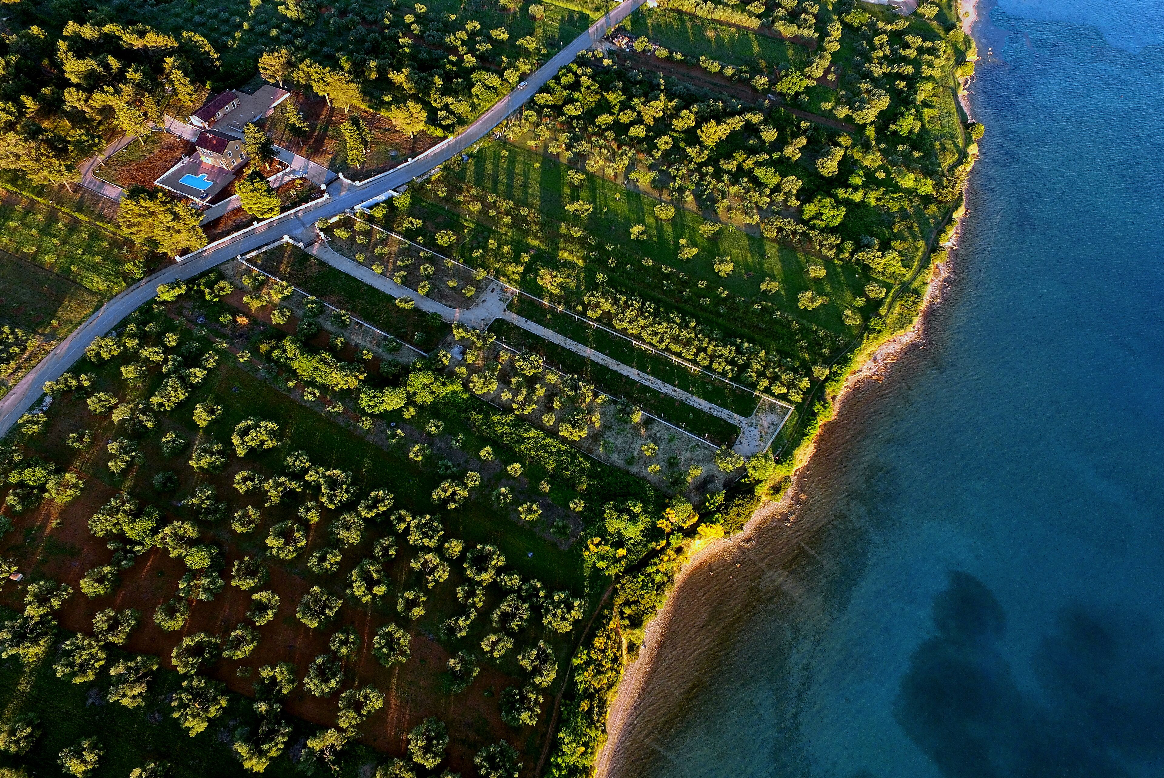 An aerial view of a coastal property with a swimming pool, a tennis court, and a road leading to the beach.