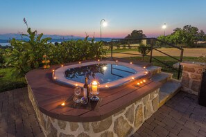 A hot tub with candles and bottles, surrounded by stone steps and greenery, overlooking a scenic landscape.