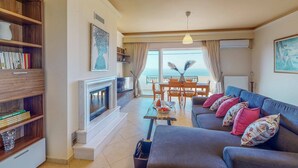 A living room with a sofa, a coffee table, and a bookshelf. There is a fireplace, a dining area with a table and chairs, and a view of the sea through large windows.