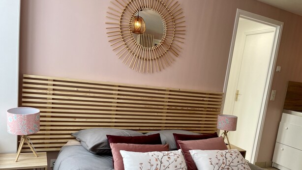 A bedroom with a wooden slat headboard, a round wall mirror, two bedside tables with lamps, and a bed with grey bedding and decorative pillows. - Le Cocon - Towncenter (Bayeux)