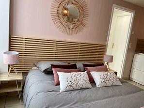 Una camera da letto con testiera in listelli di legno, uno specchio rotondo a parete, due comodini con lampade e un letto con biancheria grigia e cuscini decorativi.