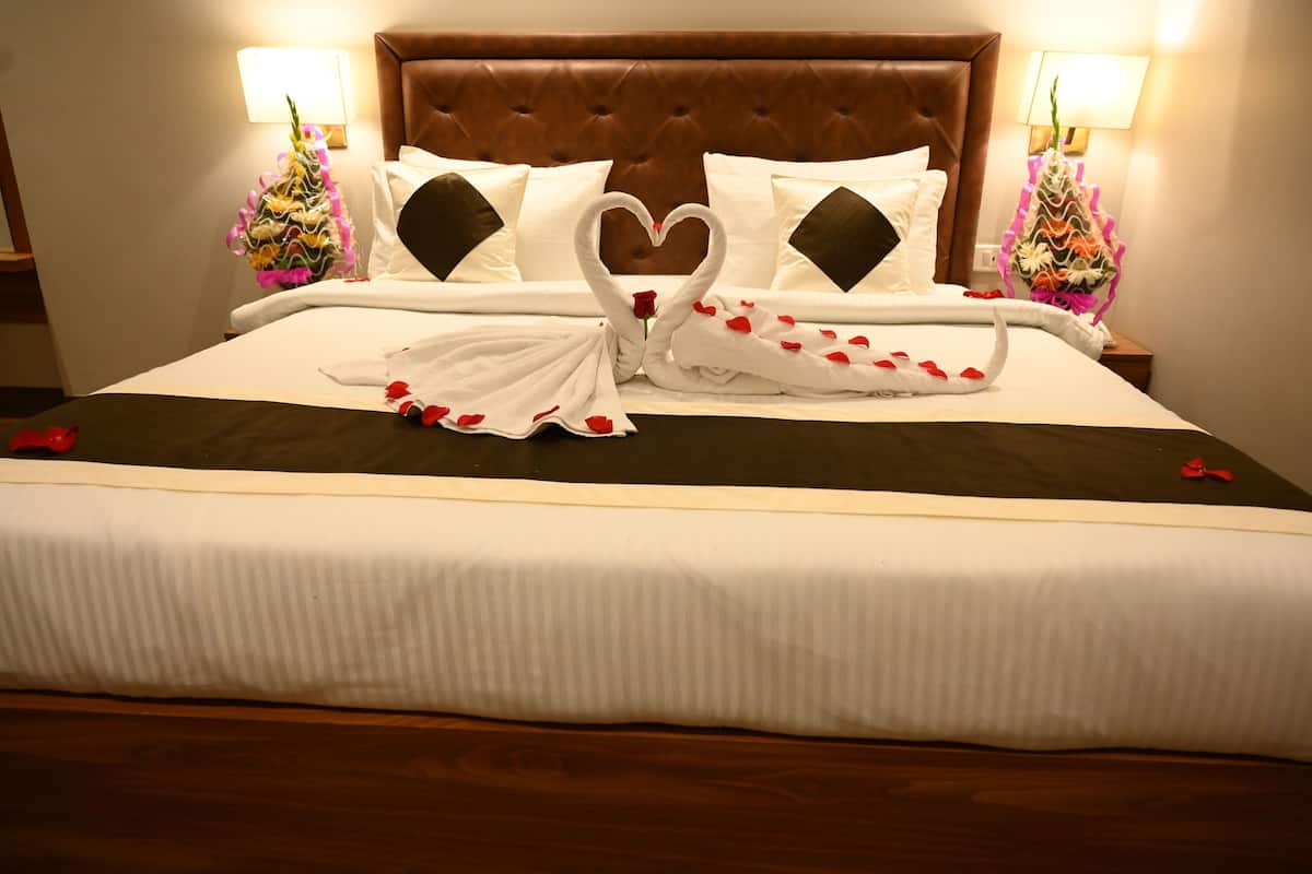 Una cama bien hecha con una toalla dispuesta en forma de corazón, un cabecero marrón y dos lámparas fijadas a la pared.
