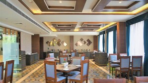 Un'area ristorazione di un hotel con diversi tavoli e sedie, una cucina sullo sfondo e piastrelle a motivi decorativi.