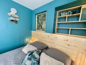 Een slaapkamer met een blauwe wand, een houten hoofdbord, een bed met grijze beddengoed, een decoratieve spiegel en een plank met boeken.
