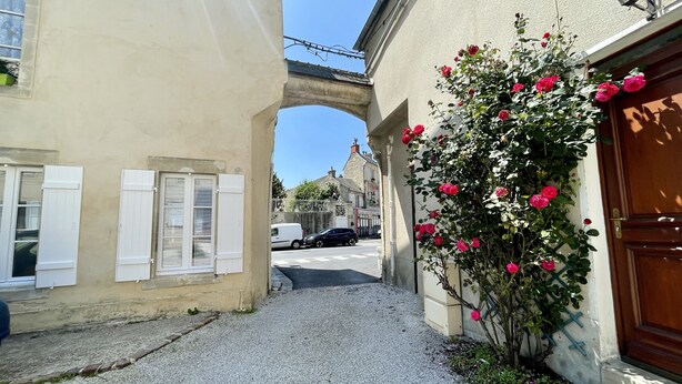 Una calle estrecha con un sendero de grava, un puente de piedra y un edificio con puerta de madera y rosal.
