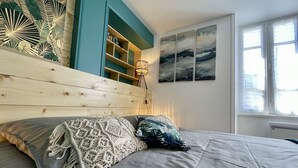 Un dormitorio con cabecera de madera, cama con ropa de cama gris, un cojín de macramé y un cojín estampado, además de una pared con arte abstracto.