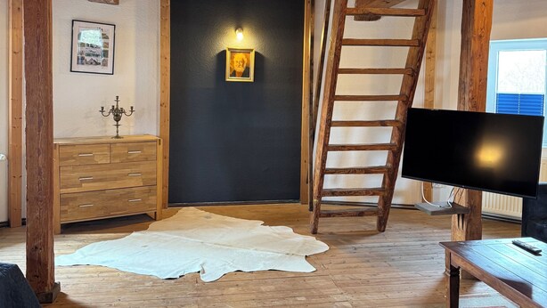 A rustic room with wooden beams, a black wall with a framed picture, a chandelier, a wooden dresser, a television, and a large fur rug.