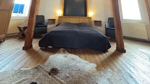 A room with a high wooden ceiling, a bed, two chairs, a window, and a radiator.