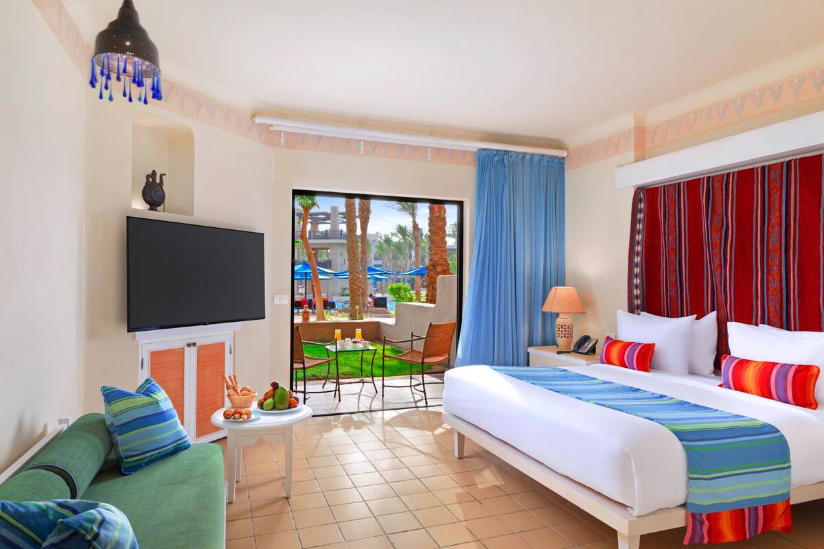 A hotel room with a bed, a TV, a small table with fruit, a sofa, and a view of the pool area.