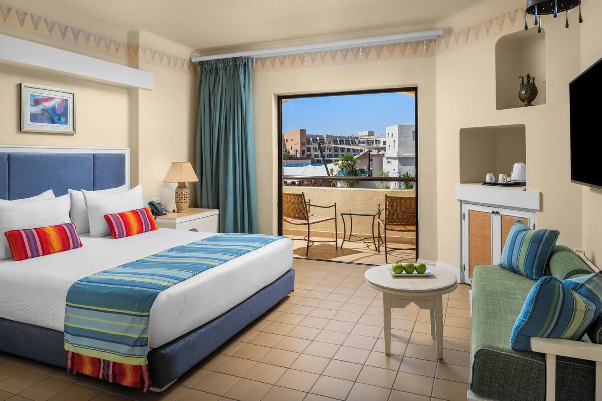 A hotel room with a bed, a sofa, a small table with fruit, a TV, and a balcony with a view of buildings.