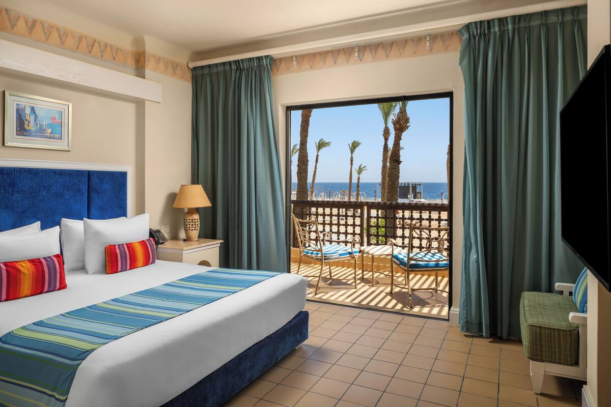 A hotel room with a large bed, a view of the sea, a balcony with chairs, and a television.