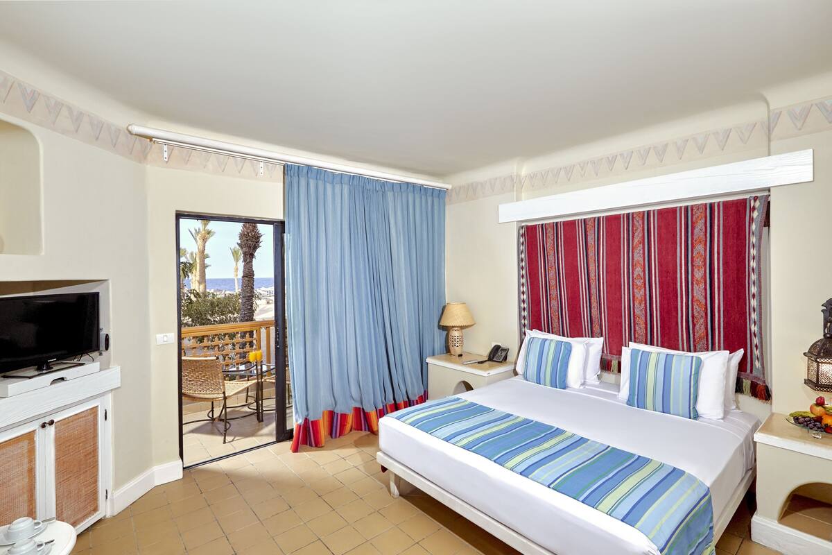 A hotel room with a bed, a TV, a balcony with a view, and a small table with a lamp.