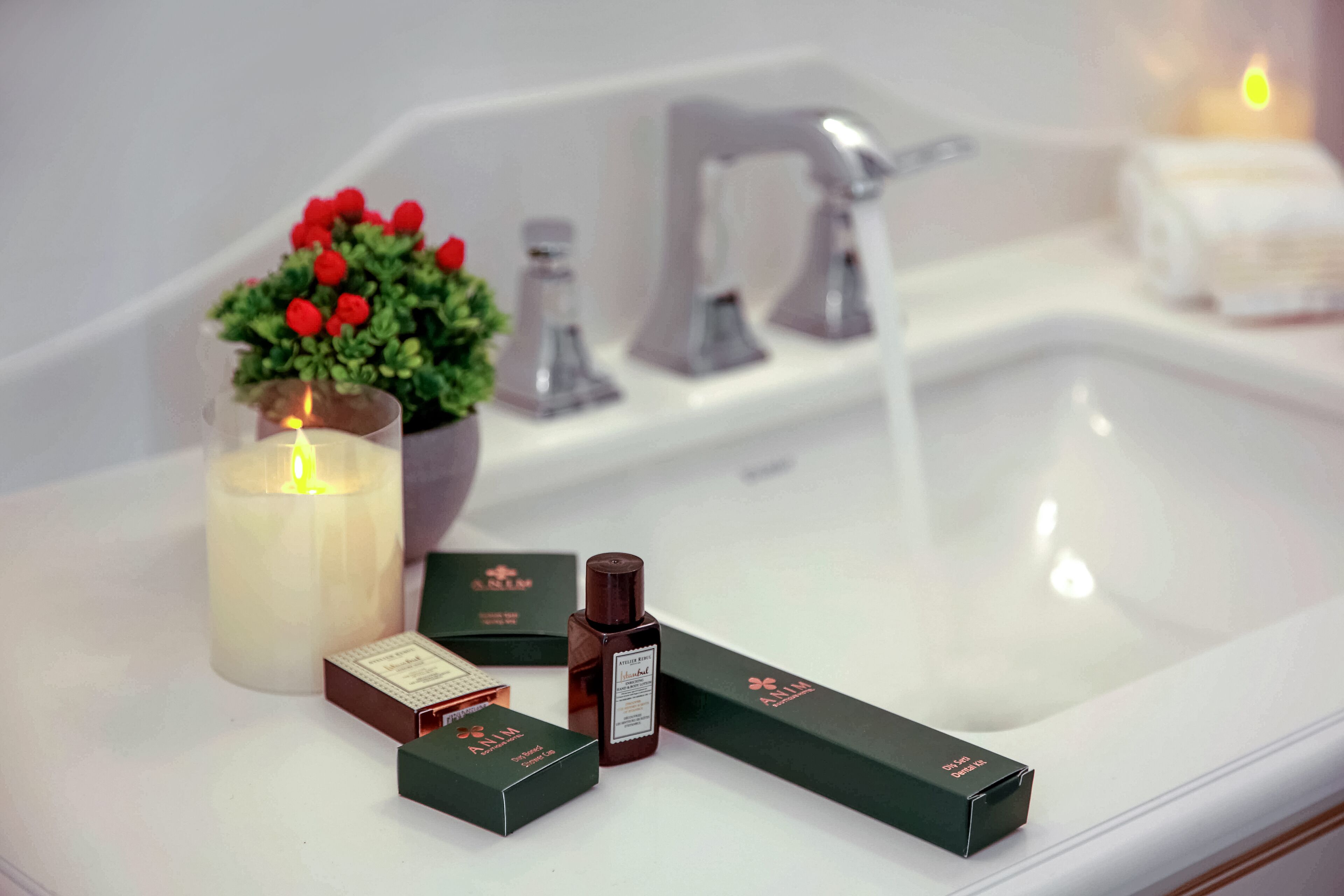 A bathroom with a white sink, a lit candle, a small potted plant with red flowers, and several skincare product boxes.