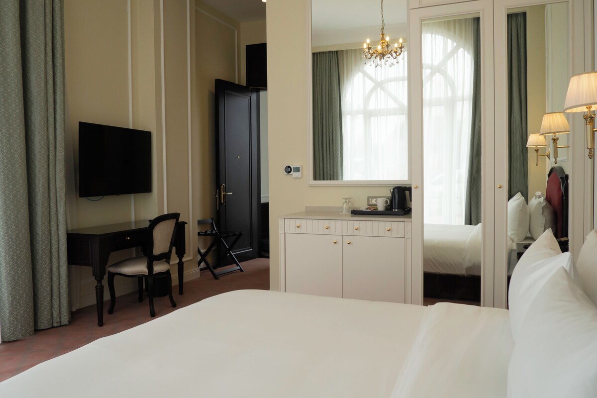 Habitación de hotel con cama, escritorio, silla, televisor, armario, espejo y candelabro.