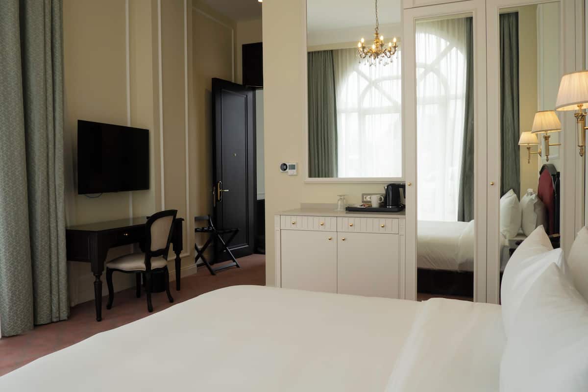 Habitación de hotel con cama, escritorio, silla, televisor, armario, espejo y candelabro.