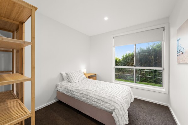 A bedroom with a bed, a nightstand, a window with a view of greenery, and built-in wooden shelving.